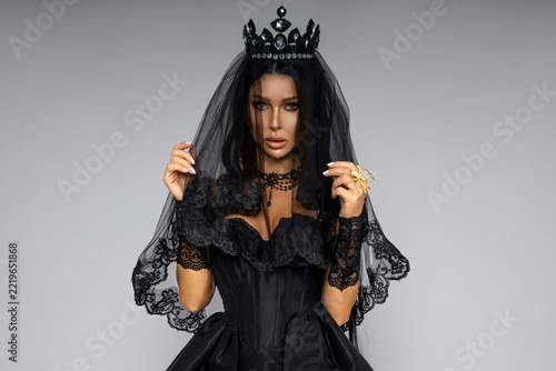 Sensual dark gothic queen in black lace veil, crown, and lingerie posing for a mysterious Halloween fashion portrait on grey background