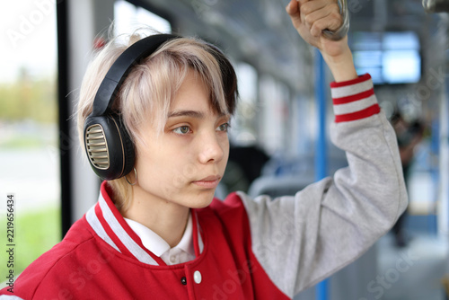 Thoughtful teenage girl rides a city tram she standing and listening to music through headphones. Urban public transport commute, technology and youth lifestyle concept.