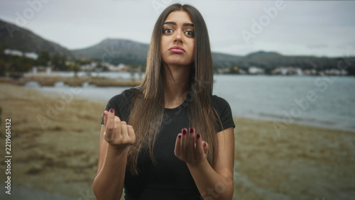 Woman with palms visible making pinched fingers and rubbing hand gesture on beach with coastal town in view; disappointment.