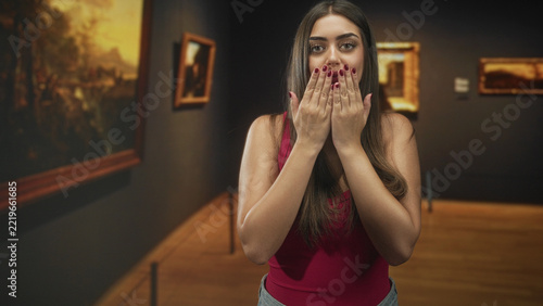 Fotografie Young woman blows a kiss with cupped hands and puckered lips toward viewer while standing in a museum gallery room with paintings and stanchions; playful affection