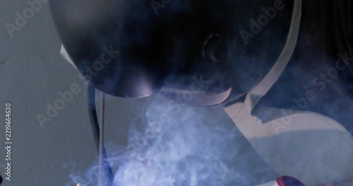 Welder in dark helmet and apron leaning over metal welding process close up