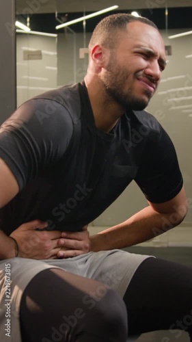 Sick African American male man athlete feeling abdominal pain in gym during abs workout suffering digestive problem belly discomfort stomach ache unwell bodybuilder guy after fitness exercise indoors