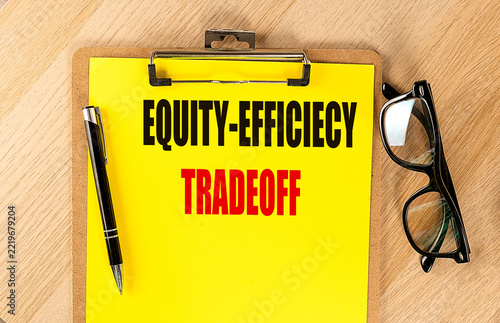Equity and efficiency tradeoff concept in economics, text yellow paper attached to a clipboard with pen and glasses on a wooden desk