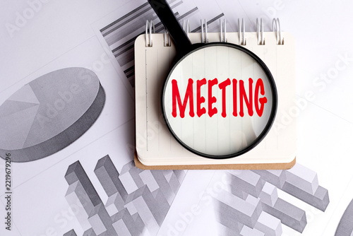 Meeting text viewed through a magnifier on a spiral notebook over business charts, planning, discussion and analysis concept