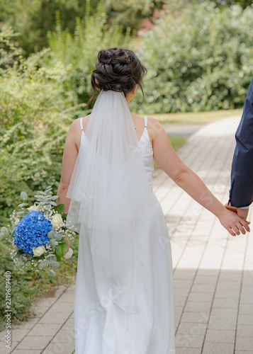romantic bride walking hand-in-hand with groom on outdoor pathway surrounded by greenery in third-person perspective from behind the bride during wedding photography session.
