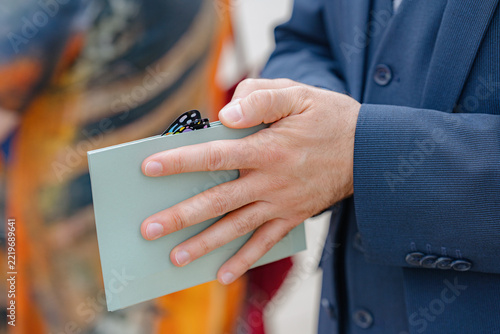 man holding wallet in formal suit at indoor event close-up focusing on hands and wallet with blue and white colors and clean background for business and fashion use cases.