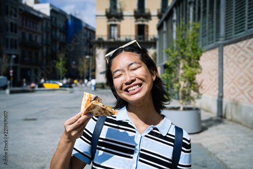 Asian woman enjoying pizza slice while smiling in city
