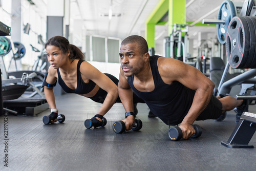 Fotografie Fit black couple performing synchronized push-ups using dumbbells as handles on