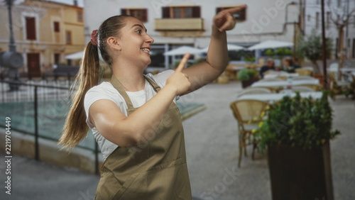 Woman in apron points thumb to herself and makes lively hand gestures in a busy street cafe setting; friendly confidence.
