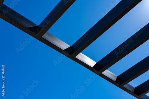Angled dark metal louvre beams crossing a clear blue sky