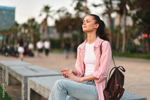 Fotografie Young woman with dark hair sits quietly on a concrete bench in a sunny park, eyes closed and mind at ease after a casual stroll
