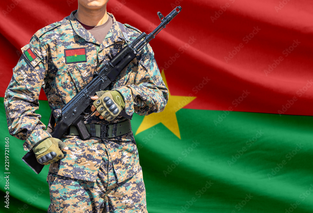 Fototapeta premium Burkina Faso flag with soldier in military uniform holding assault rifle in front of it