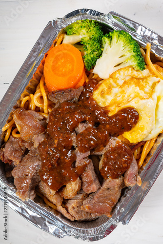Beef noodles with fried egg and fresh vegetables packed in aluminum foil takeout tray