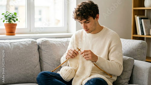Young man sits on a sofa carefully stitching a soft fabric object by hand concentrating on his needlework and enjoying a peaceful creative home craft session