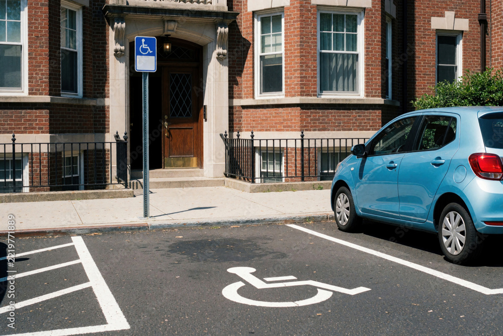 Fototapeta premium Blue car parked in accessible parking space near brick residential building entrance. Handicap symbol painted on asphalt with blue sign on pole. Disabled parking lot allows easy access for people