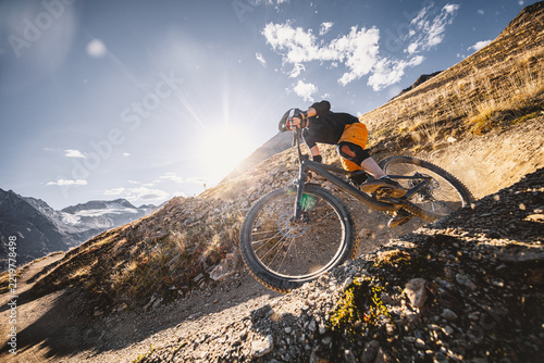 Mountain biker riding down a steep rocky alpine trail with dramatic mountains and a turquoise lake below, captured in warm sunlight during an adventurous high altitude descent ride.