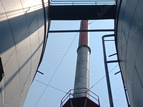 Industrial Chimney Tower and Metal Storage Tanks Under Clear Blue Sky.