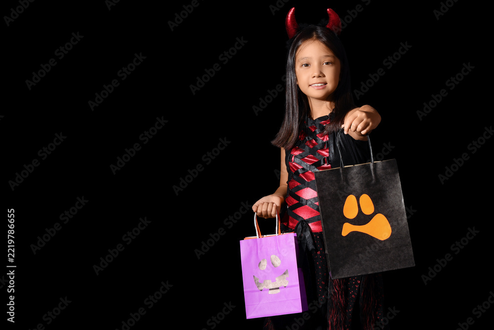 Fototapeta premium Halloween trick or treat, happy girl in devil costume holding candy bags for holiday celebration and autumn festivities on a black background