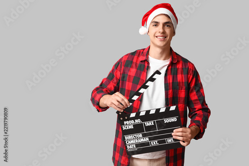 Young man in Santa hat with clapperboard on grey background. Christmas celebr...