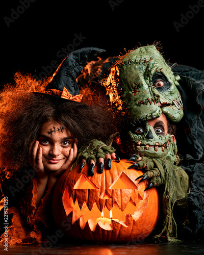 A playful Halloween scene featuring a girl in a witch hat and a child dressed as Frankenstein's monster behind a glowing carved pumpkin, with dramatic orange lighting and a dark background.