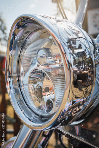 Close-up of a chrome motorcycle headlight with engraved details, reflecting the surrounding outdoor scene.