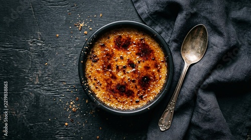 Classic French creme brulee with a burnt caramelized sugar crust rests in a black ceramic ramekin beside a silver spoon in a top down view