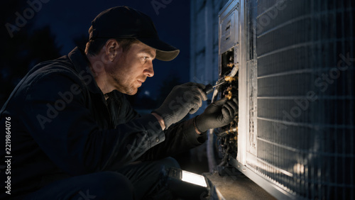 HVAC maintenance worker repairing outdoor cooling equipment at night under focused work light