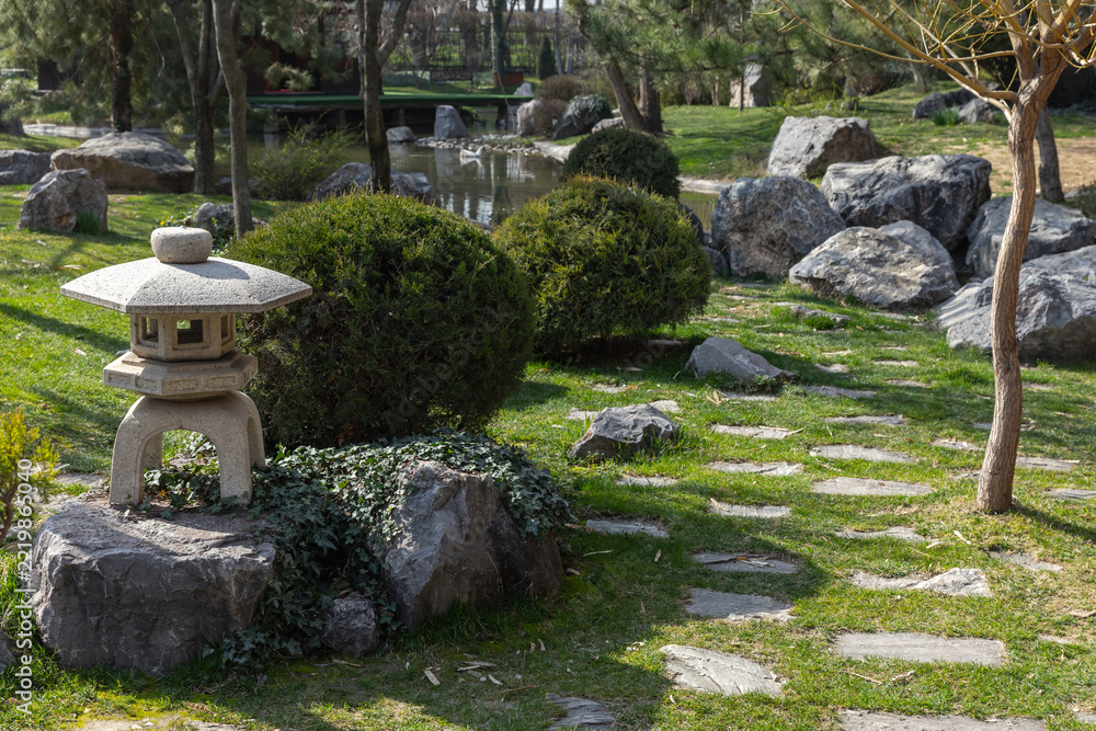 Fototapeta premium A serene Japanese garden scene featuring a traditional stone lantern beside stepping stones, pond reflections, rounded shrubs and natural rocks, evoking tranquility, meditation and landscape design