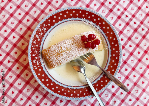 A slice of traditional Bavarian quark strudel dusted with powdered sugar, served with creamy sauce and red currants on a decorative red plate