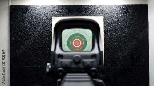 Red holographic reticle sight aimed at green paper target at indoor shooting range, precision aiming and marksmanship training concept