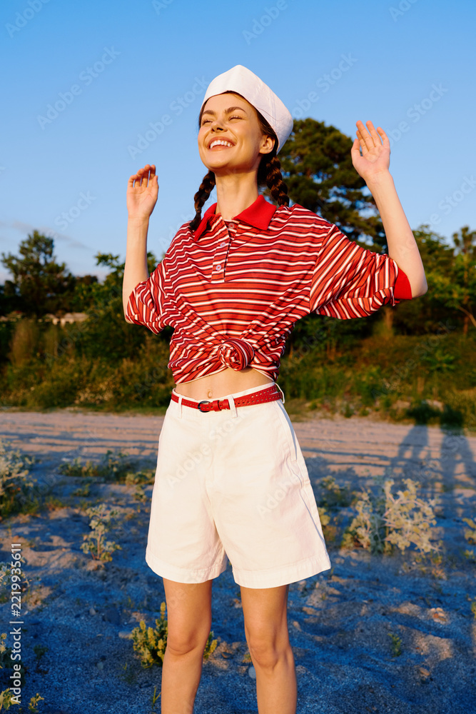 Naklejka premium A happy young woman in a striped shirt and shorts poses outdoors enjoying summer, lifestyle concept with casual clothes on a sandy beach during sunset, celebrating freedom and joy daily.