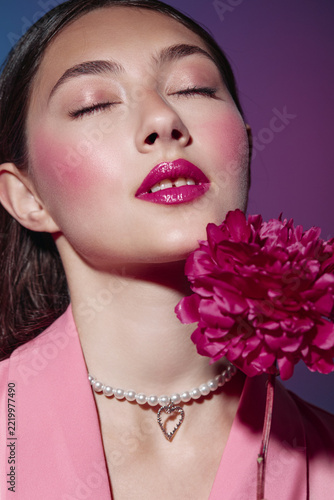 Close-up of a young woman with glossy fuchsia lips and pink blush. She wears a pearl necklace and pink blazer while holding a matching vibrant flower. Fashion beauty concept.