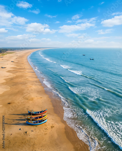 Aerial view of colorful fishing boats resting on a sandy beach beside calm ocean waves under a cloudy blue sky