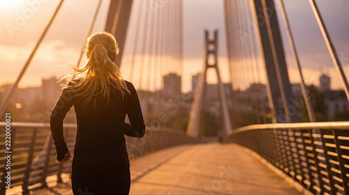 Runner Woman Exercising on a Modern Pedestrian Bridge at Sunset with City Silhouette Backdrop for Healthy Lifestyle and Urban Fitness Concept