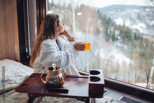 Drinking hot tea enjoying winter landscape in mountains. Woman wears bathrobe relaxing by big window of cabin house on holiday