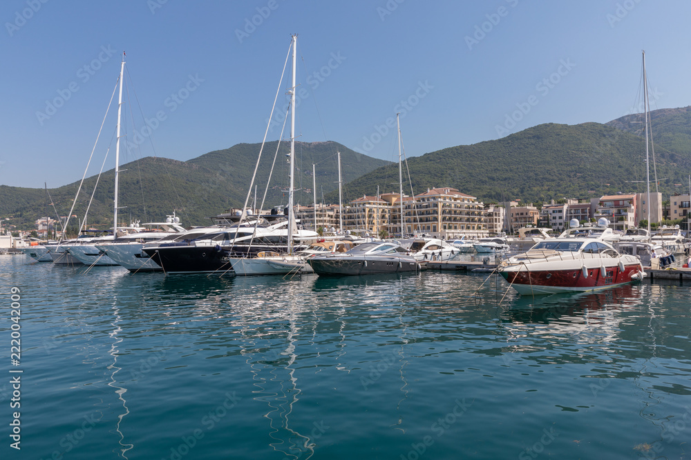 Obraz premium Tivat, Montenegro - August 11, 2025: Moored luxury yachts and motorboats rest on calm blue sea waters along coastal resort pier in Tivat, surrounded by mountain slopes and modern harbor architecture