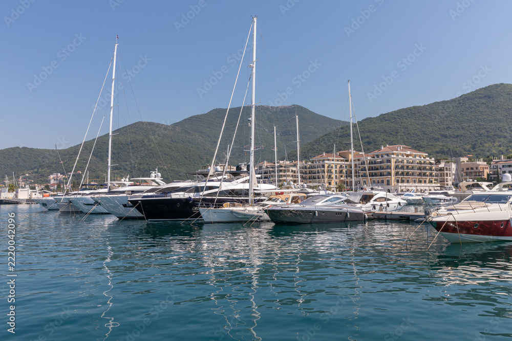 Obraz premium Tivat, Montenegro - August 11, 2025: Luxury yachts and motorboats rest in calm harbor waters of Tivat, Montenegro, featuring coastal architecture and lush green hills under bright summer sky