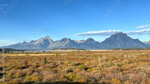 Golden grasses stretch toward jagged peaks of Grand Tetons mountain range. Blue sky frames distant mountain ridgelines. Sunlight bathes earthy tones in warm glow. No people disturb the vast, silent ex