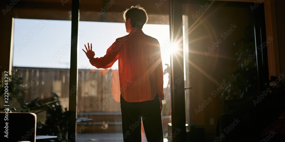 Fototapeta premium Silhouette of a person at a glass door with strong sunburst and warm light filling a cozy interior during sunrise