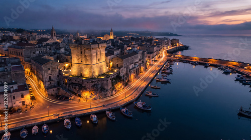 Stunning aerial view of the historic harbor of Giovinazzo in Puglia, Italy, featuring an illuminated medieval tower at sunset with boats in the calm water.