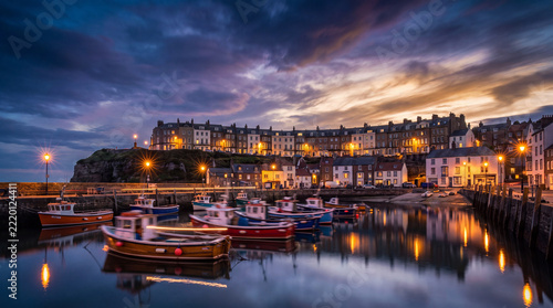 A scenic evening view of Whitby harbor in North Yorkshire, England, featuring moored fishing boats reflecting in the water under a dramatic twilight sky.