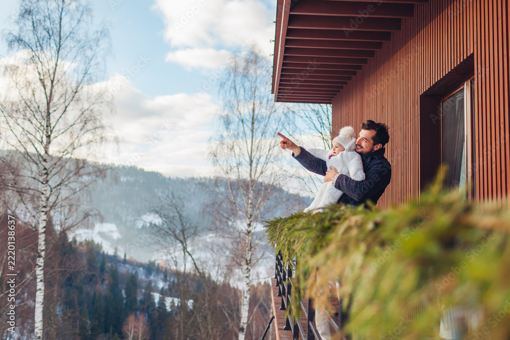 Fototapeta premium Dad and his baby girl enjoy snowy mountain landscape from hotel terrace. Family spends weekend in rented house for winter holidays