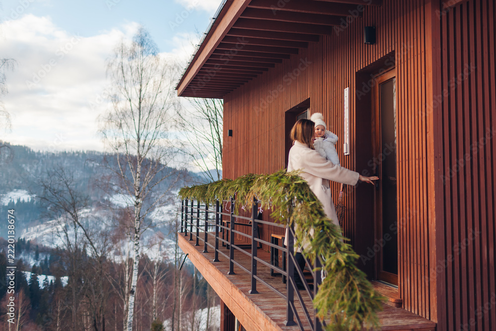Fototapeta premium Happy family renting cabin in snowy mountains for Christmas. Woman enters house decorated for holidays holding toddler baby girl