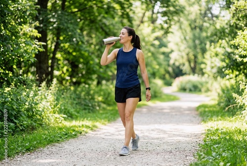 Person drinking water while walking in a sunny park. Bright daylight style for cardiovascular health, active lifestyle, and thrombosis prevention.