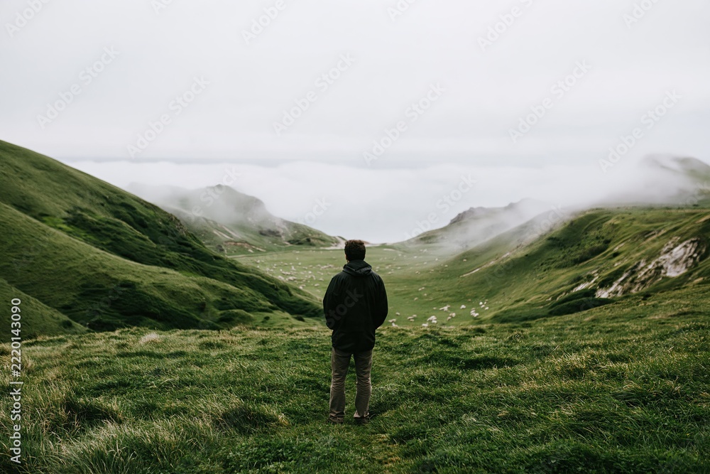 Fototapeta premium A lone figure stands on a cliffside at dawn overlooking a distant valley and mountains while clouds slowly drift across the landscape below