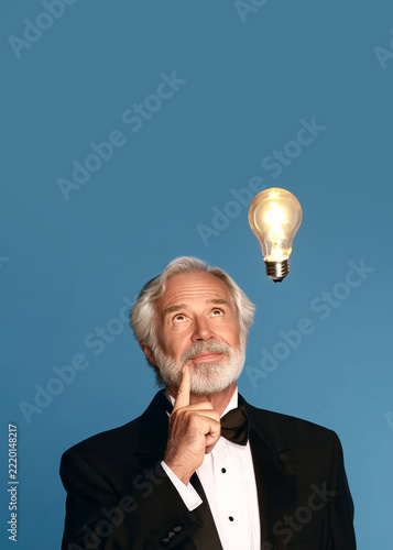 Elderly Gentleman with White Beard in Formal Tuxedo Gazing at Glowing Lightbulb Above His Head, Symbolizing Sudden Idea and Inspiration, Stark Blue Background, Vertical 5x7, Copy Space