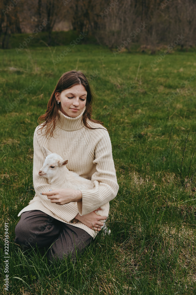 Naklejka premium Bright outdoor scene with a woman in a cozy cream sweater sitting in a grassy field, gently holding a white goat kid, capturing a peaceful, tender lifestyle moment in sunlight