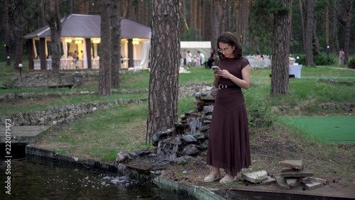 Young woman standing by an artificial waterfall with a phone. Girl wearing glasses and a dress stands beside a decorative waterfall with a festive tent in background.