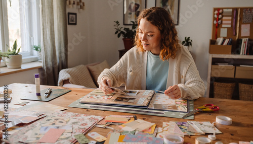 A creative woman joyfully curates a personalized scrapbook at her sunlit wooden table. This cozy scene captures the nostalgic art of preserving memories, ideal for lifestyle or hobby projects.