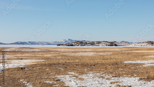 Panoramic view of a vast winter steppe landscape with dry yellow grass and snow patches leading to frozen Lake Baikal in Irkutsk Oblast, Russia under a clear blue sky on a cold sunny day